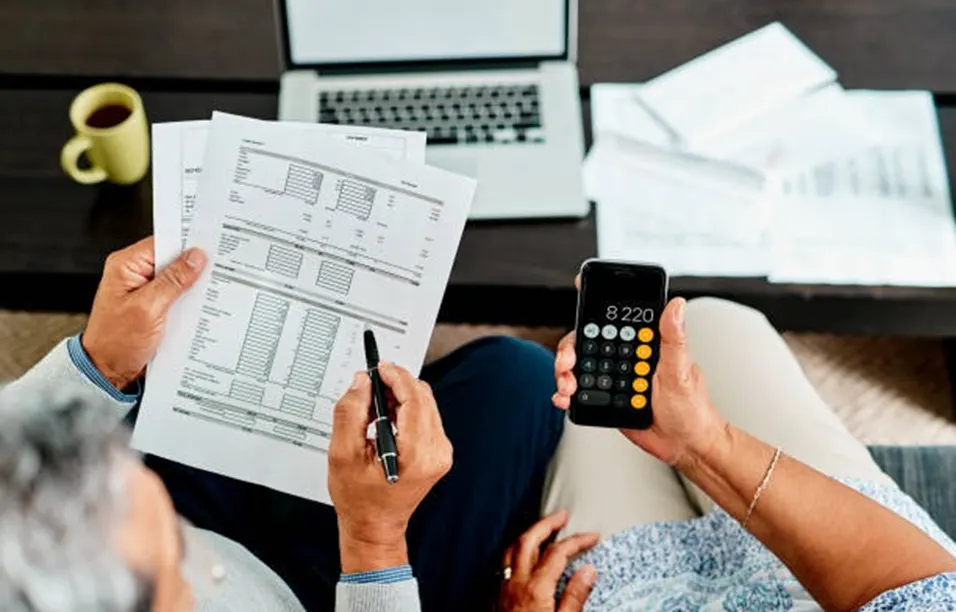 Older couple reviewing financial documents and using a phone calculator at home with a laptop nearby