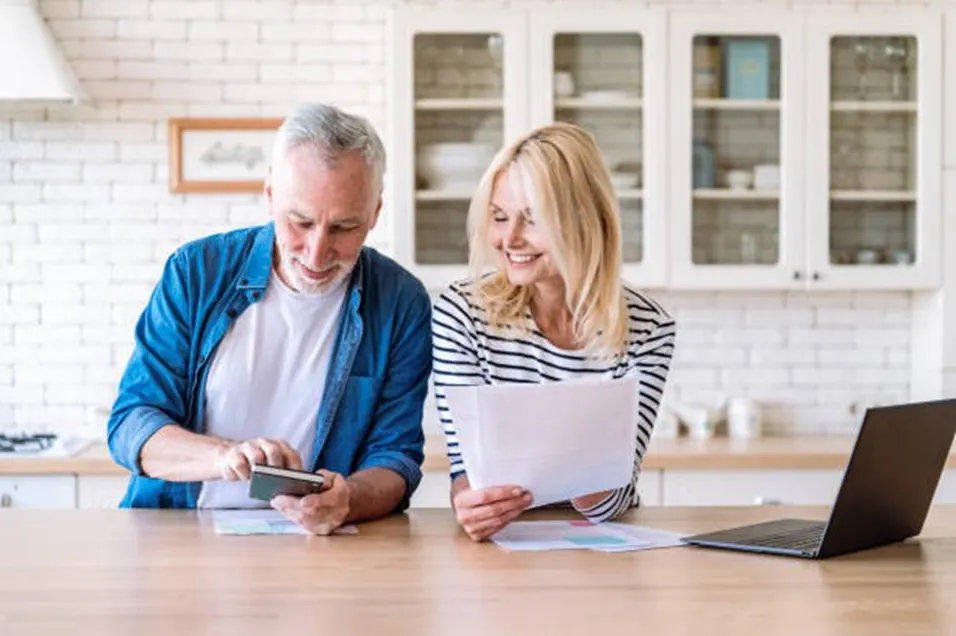 Smiling older couple using a smartphone calculator and reviewing paperwork in a bright kitchen