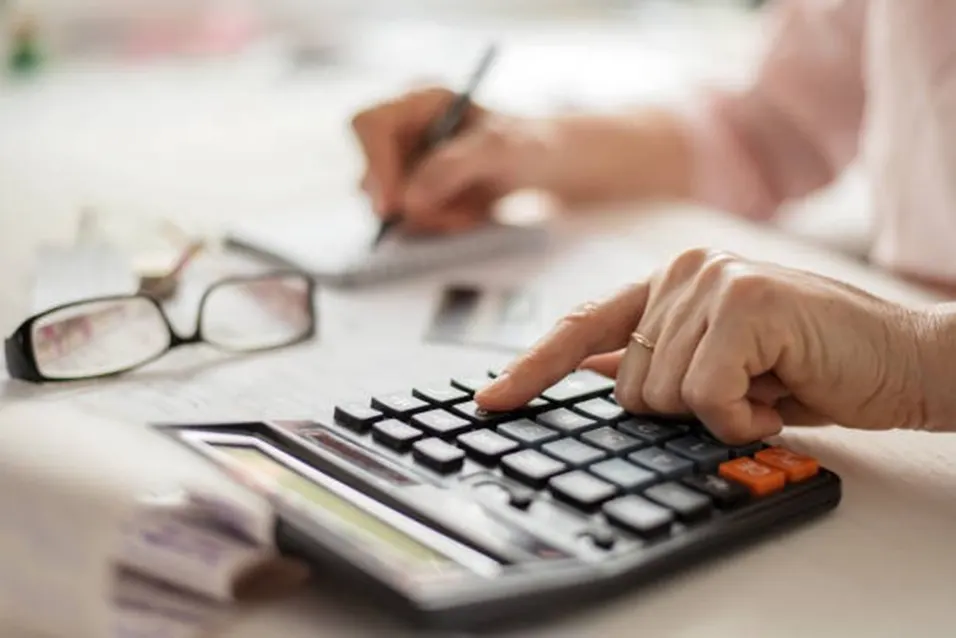 Older person using a calculator and writing notes at a desk with glasses nearby