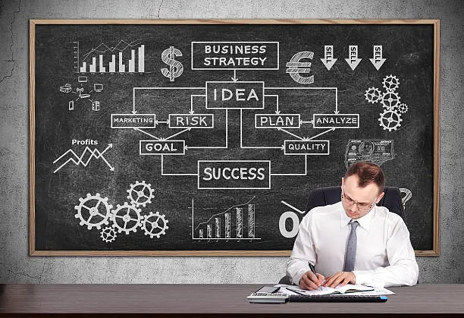 Businessman writing at a desk in front of a chalkboard showing a business strategy flowchart