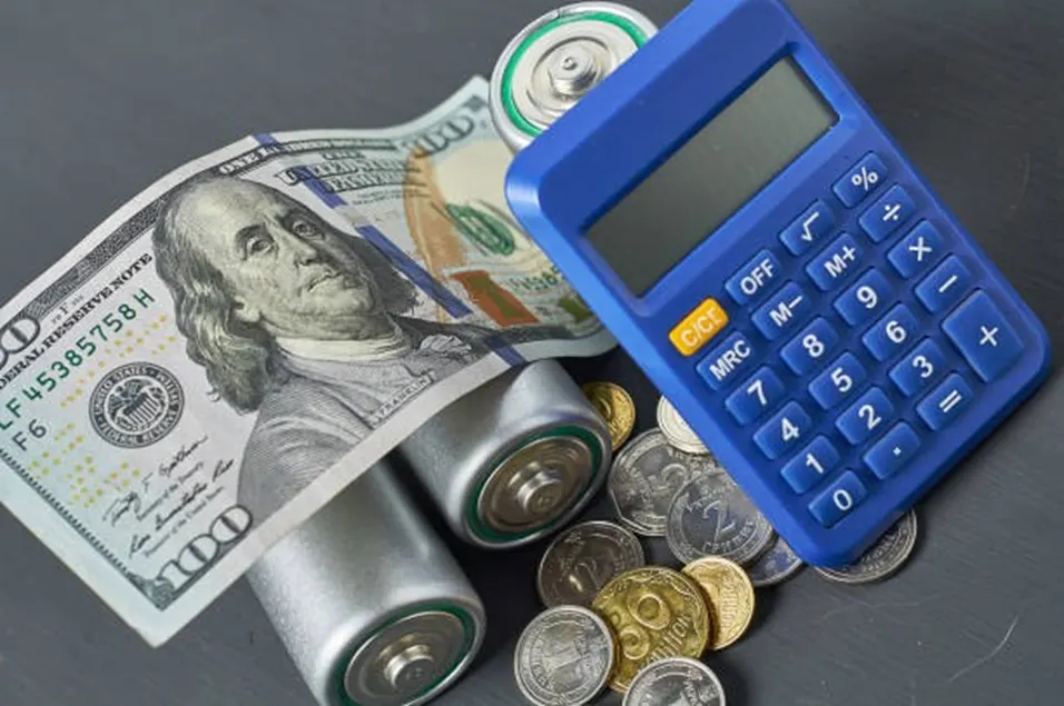 Blue calculator with US dollar bills, coins, and batteries scattered on a dark surface