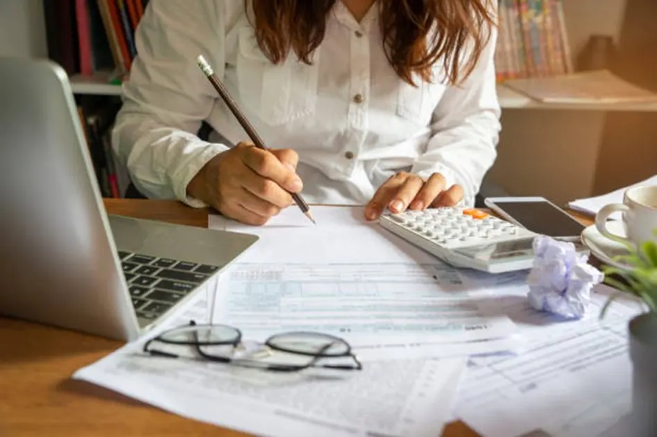 Woman writing on forms and using a calculator at a cluttered desk with a laptop and glasses