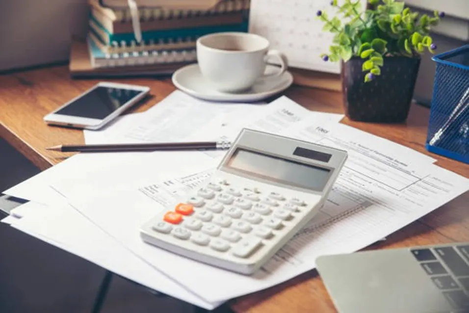 Calculator on top of tax documents with a pencil, phone, coffee cup and plant on a wooden desk