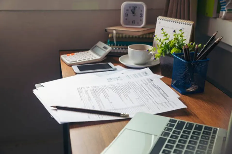 Wooden desk with tax forms, a pencil, calculator, phone, coffee cup, calendar, plant and pencil holder