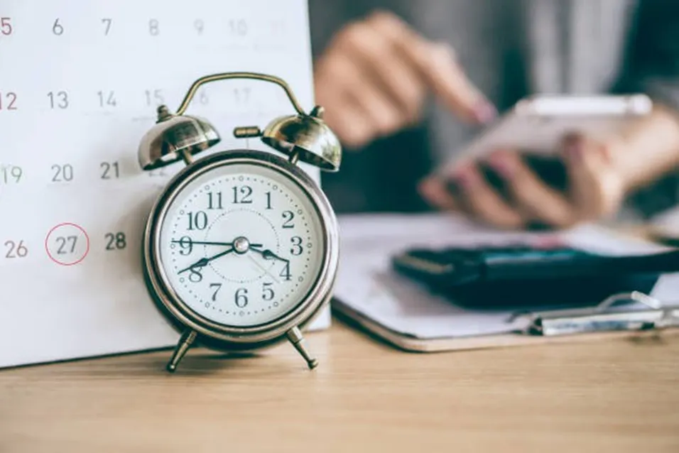 Vintage alarm clock in front of a calendar with day 27 circled, and a person using a calculator in the background