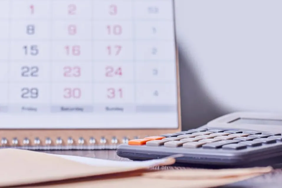 Wall calendar showing dates 1 to 31 beside a dark calculator with an orange button on a wooden surface