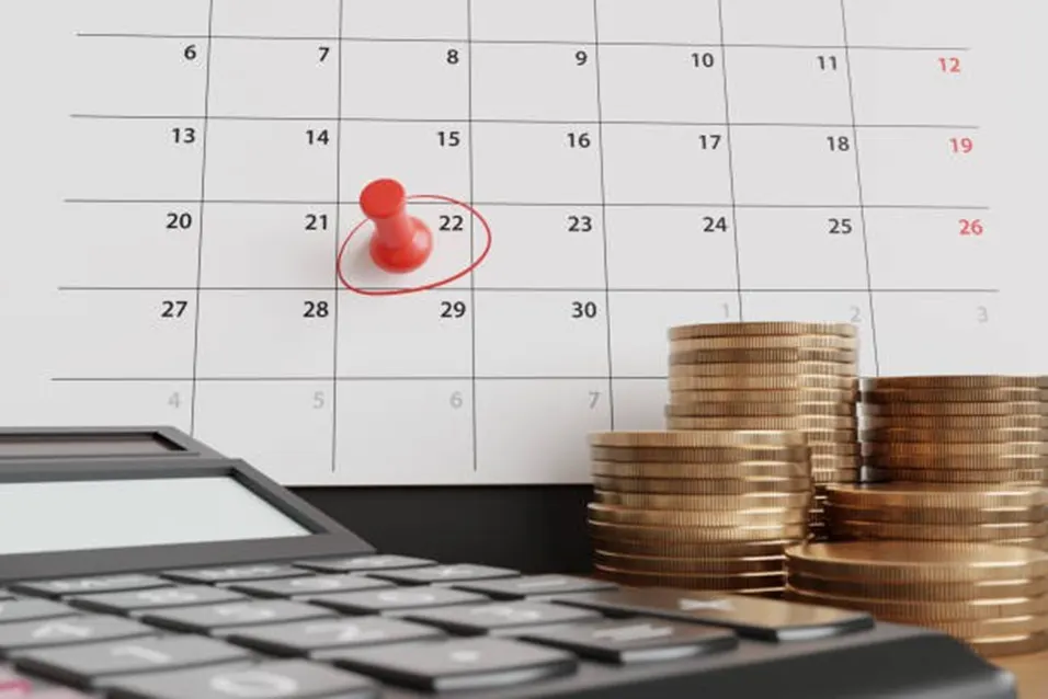 Calendar with a red pin on the 22nd beside stacks of gold coins and a black calculator