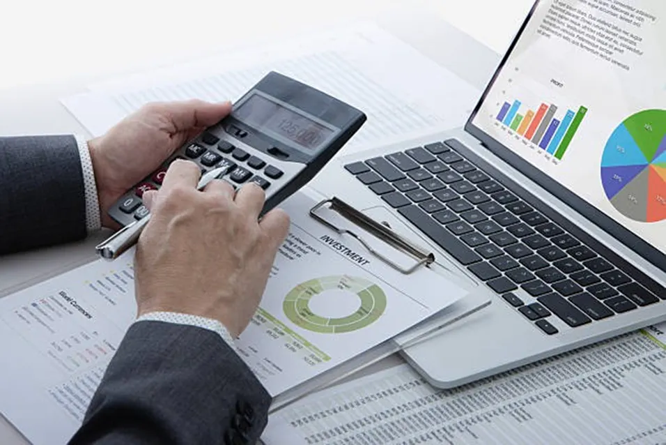 Businessman using a handheld calculator beside investment reports and a laptop showing charts