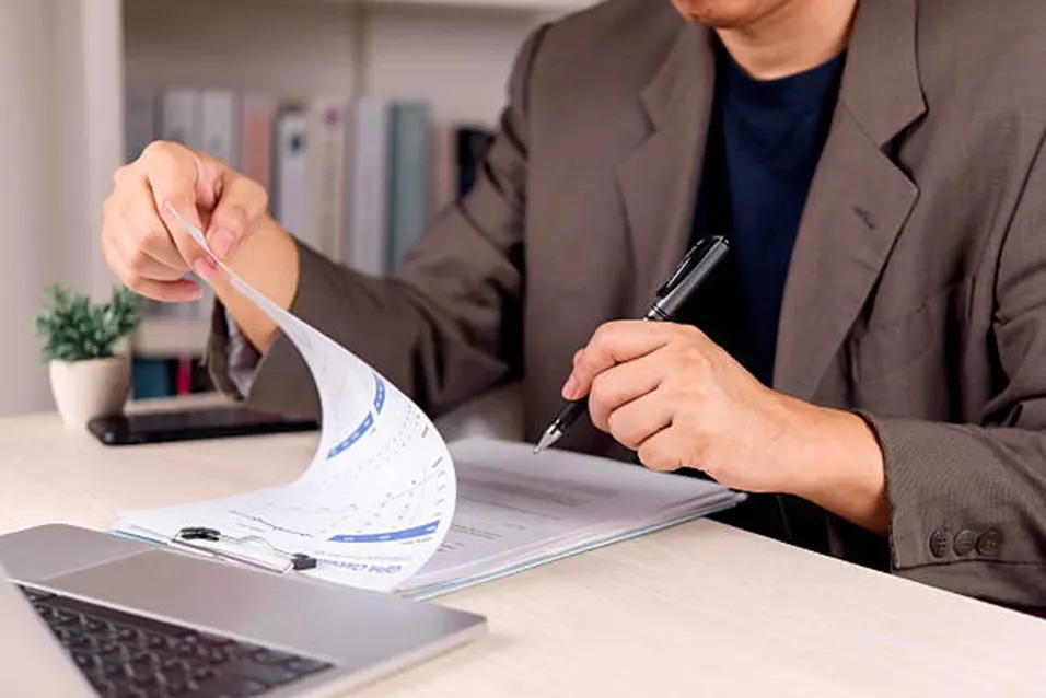 Person in a suit flipping through documents on a clipboard at a desk with a laptop