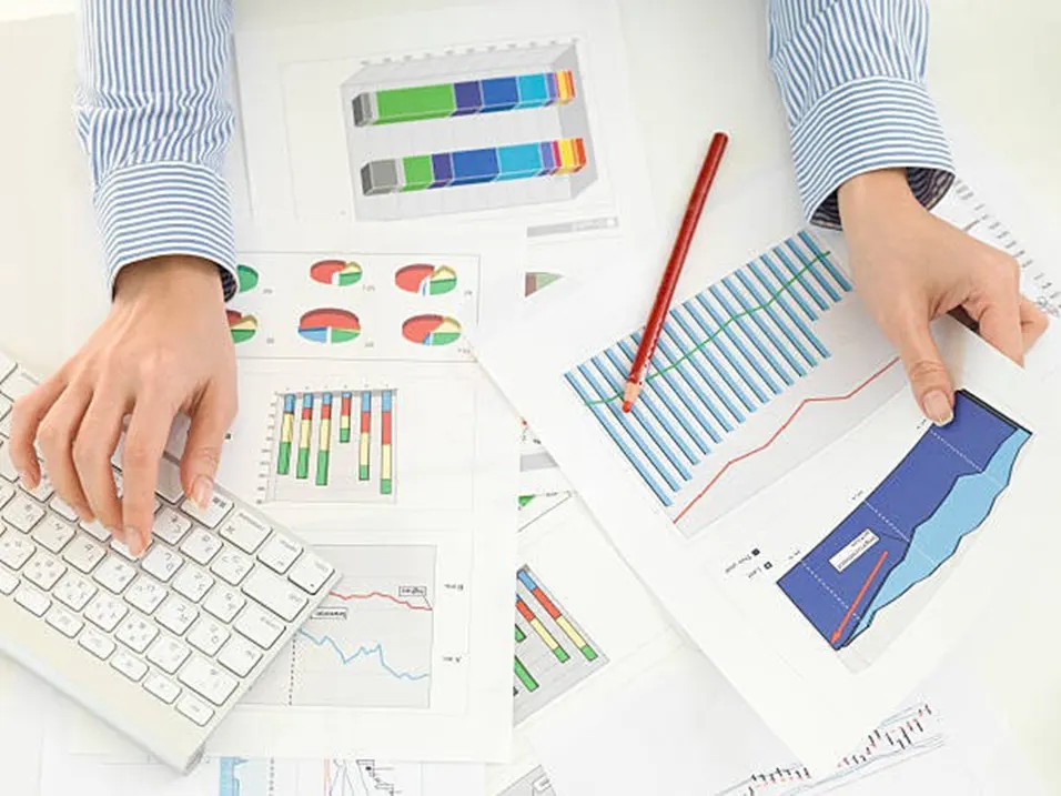 Overhead view of hands reviewing printed bar charts and pie charts on a desk with a keyboard and red pencil