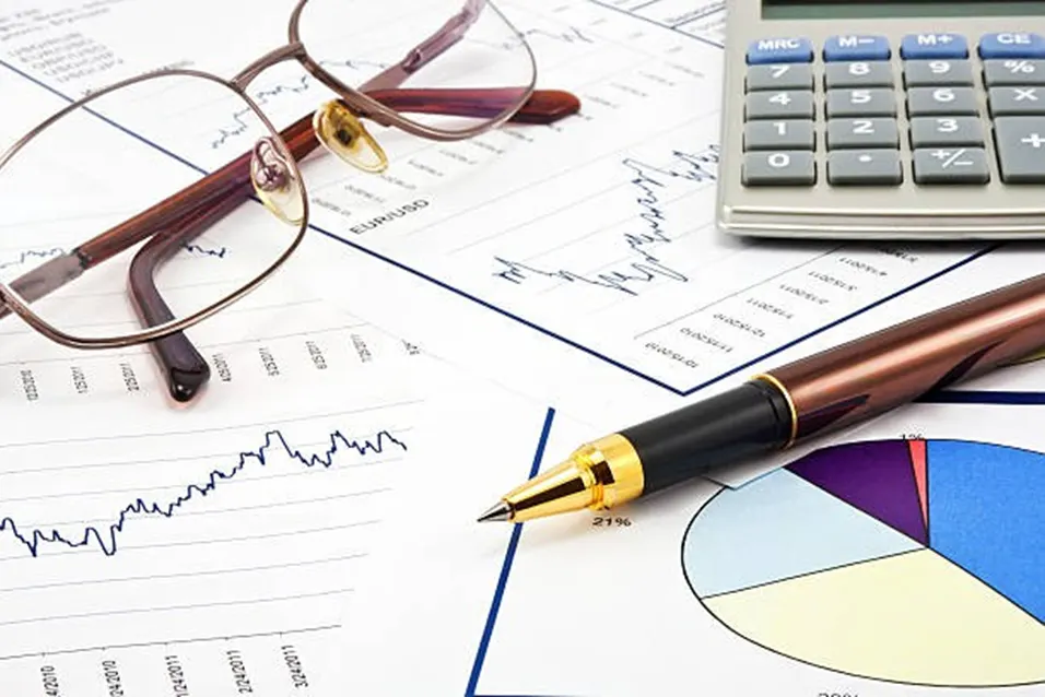 Gold-tipped pen and reading glasses resting on printed financial charts beside a calculator on a white surface