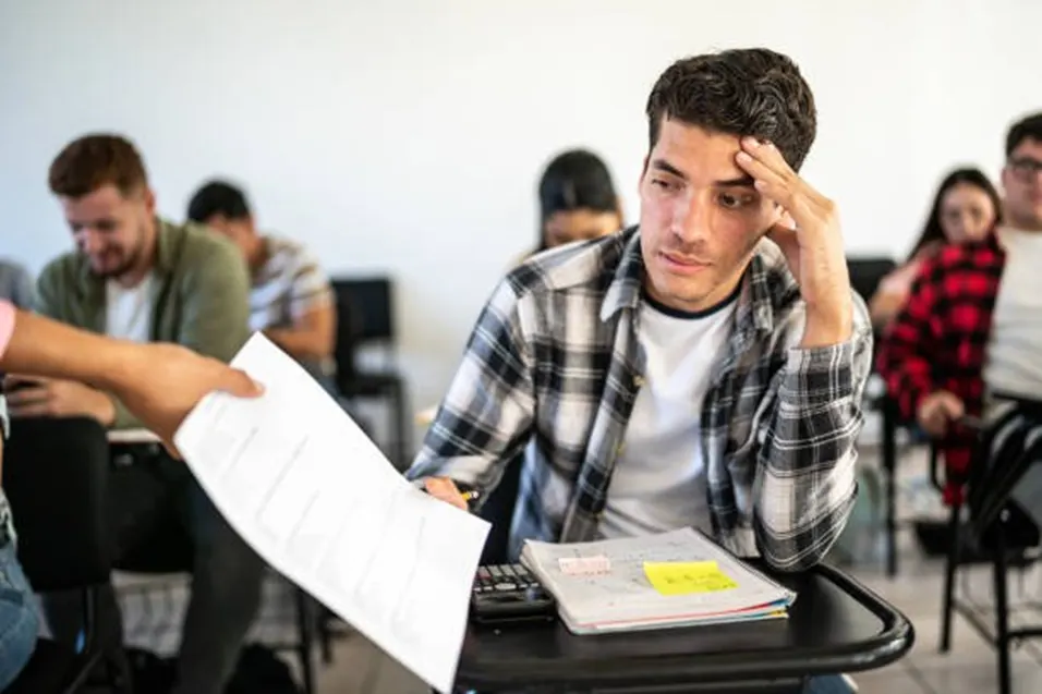 Student in a plaid shirt looking at a test paper being handed to him in a busy classroom