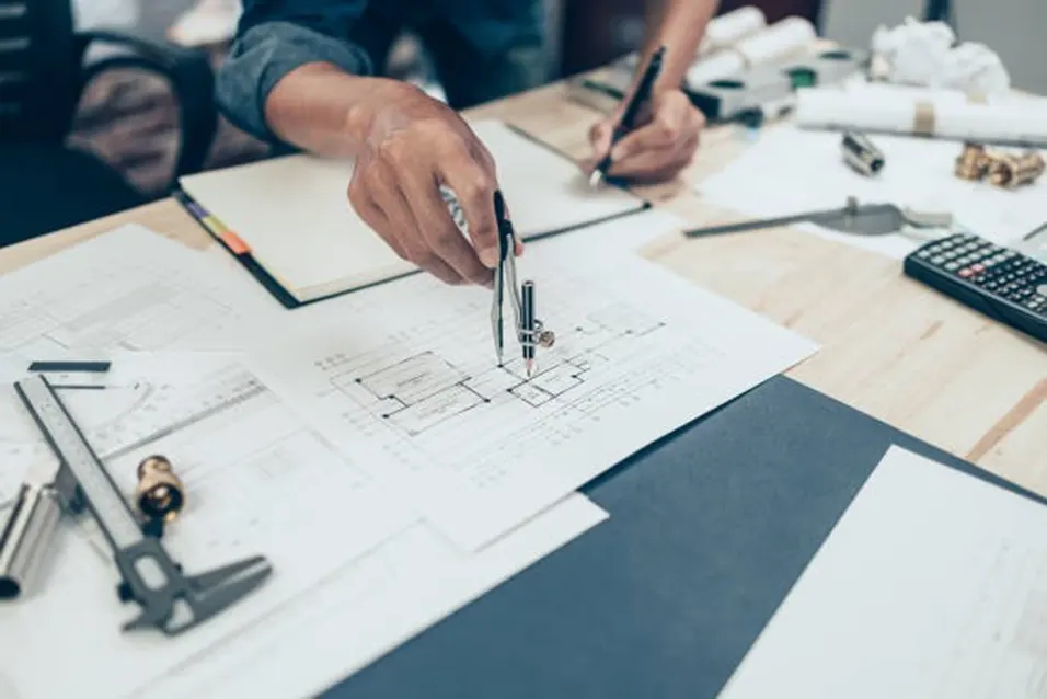 Engineer using a compass on technical blueprints at a wooden desk with a calculator and drafting tools