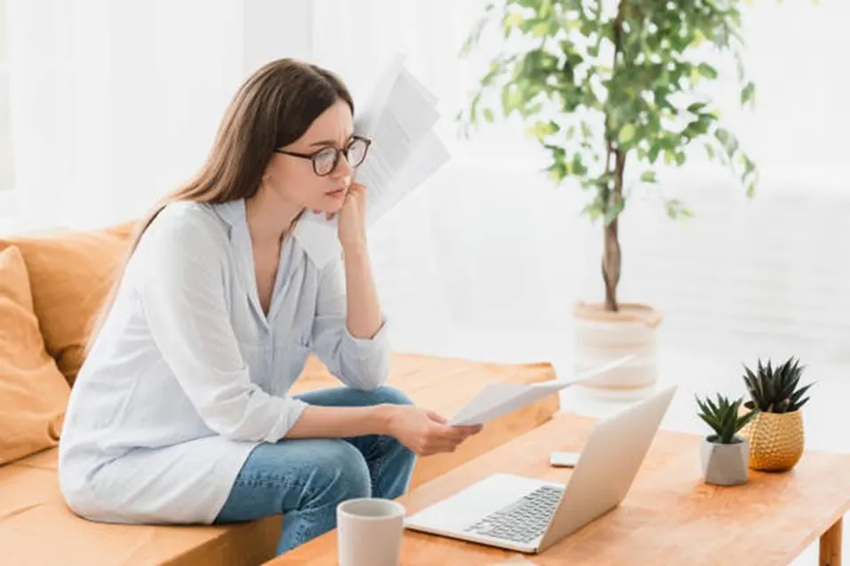 Woman with glasses sitting on a sofa reading a document beside a laptop and houseplants