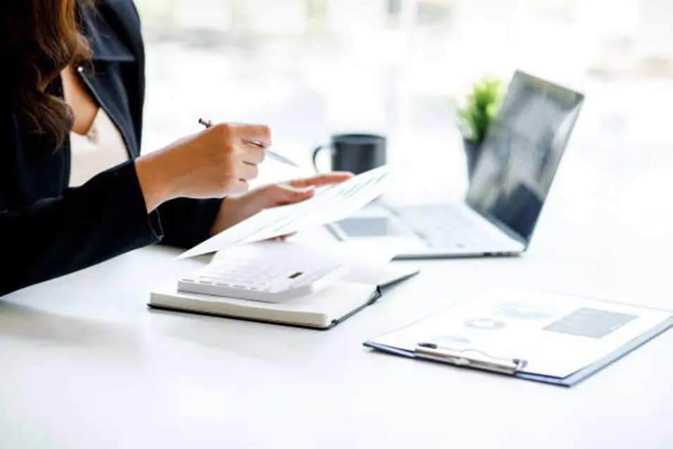 Businesswoman in a dark blazer reviewing documents with a pen and white calculator on a bright office desk