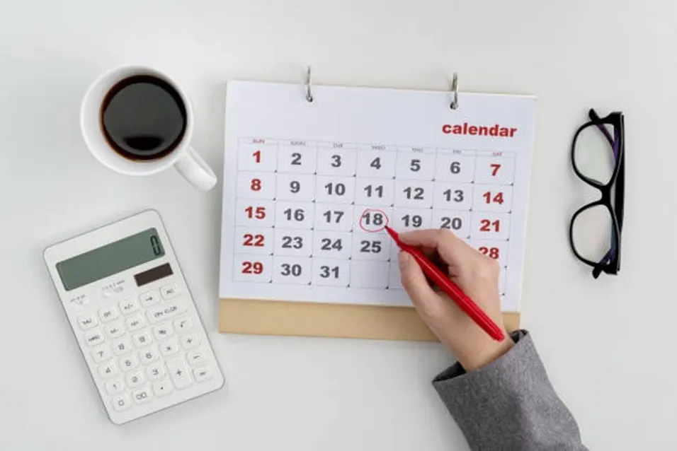Hand circling a date on a desk calendar beside a calculator, coffee and glasses on a white background