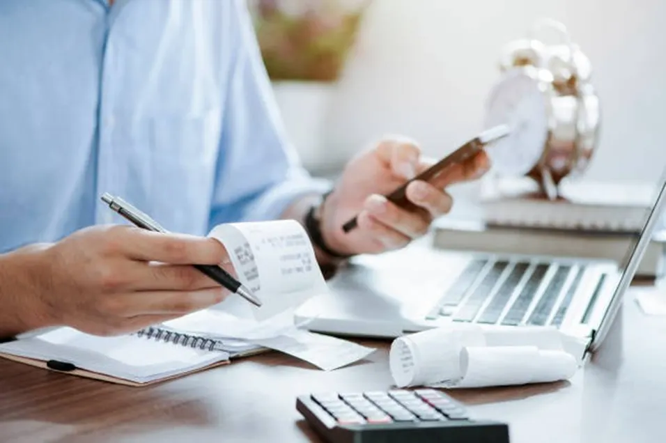 Man in a blue shirt reviewing a receipt roll and using a phone at a desk with a calculator, notebook and alarm clock