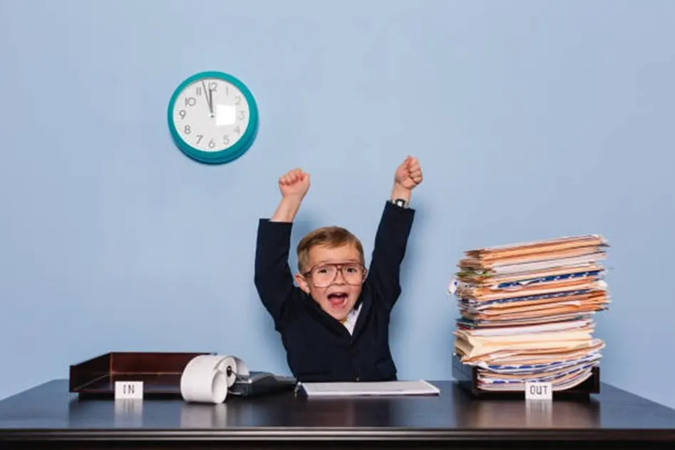 Young boy in a suit and glasses raising both arms in celebration at a desk with a clock, in-tray and stack of files