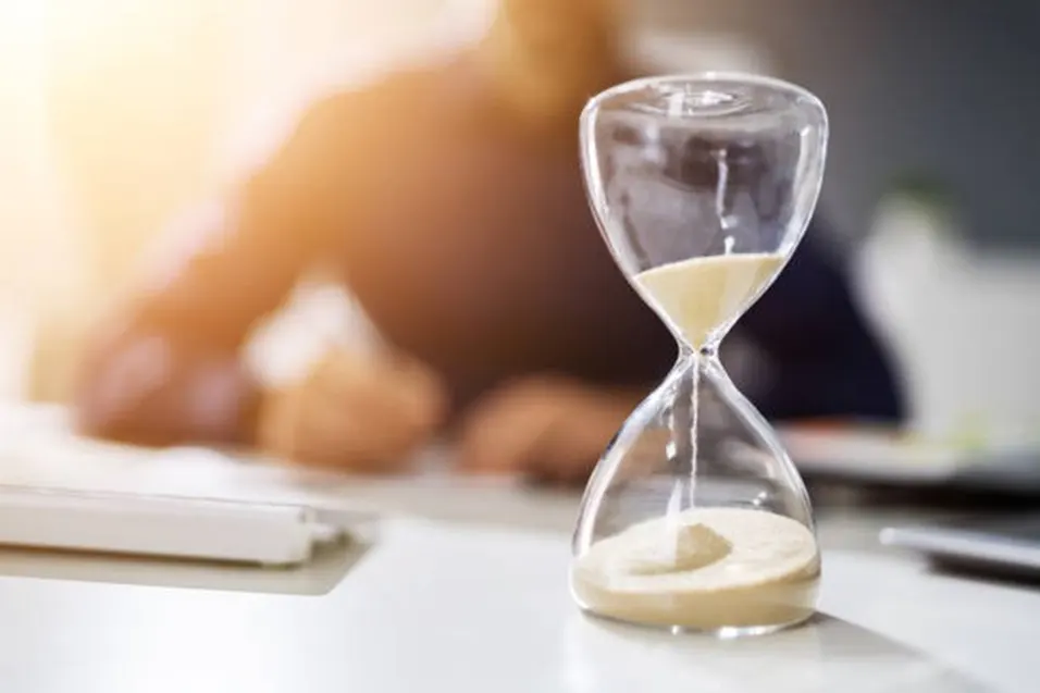 Hourglass with sand flowing on a bright white desk with a person working in the background