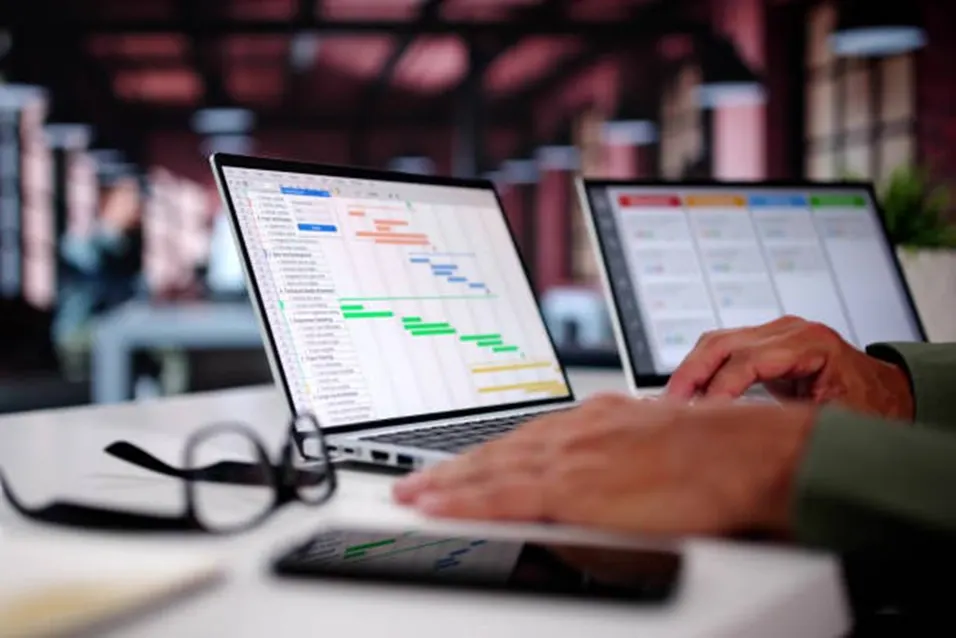 Person typing on a laptop showing a colourful Gantt chart, with a second monitor and glasses nearby in an office