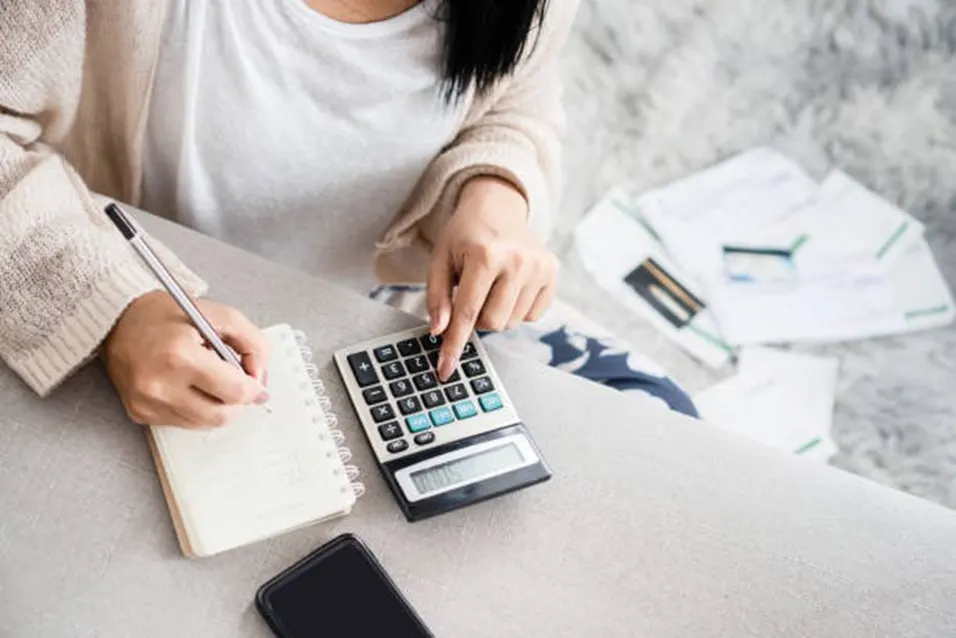 Woman using a calculator and writing notes while reviewing financial documents at home