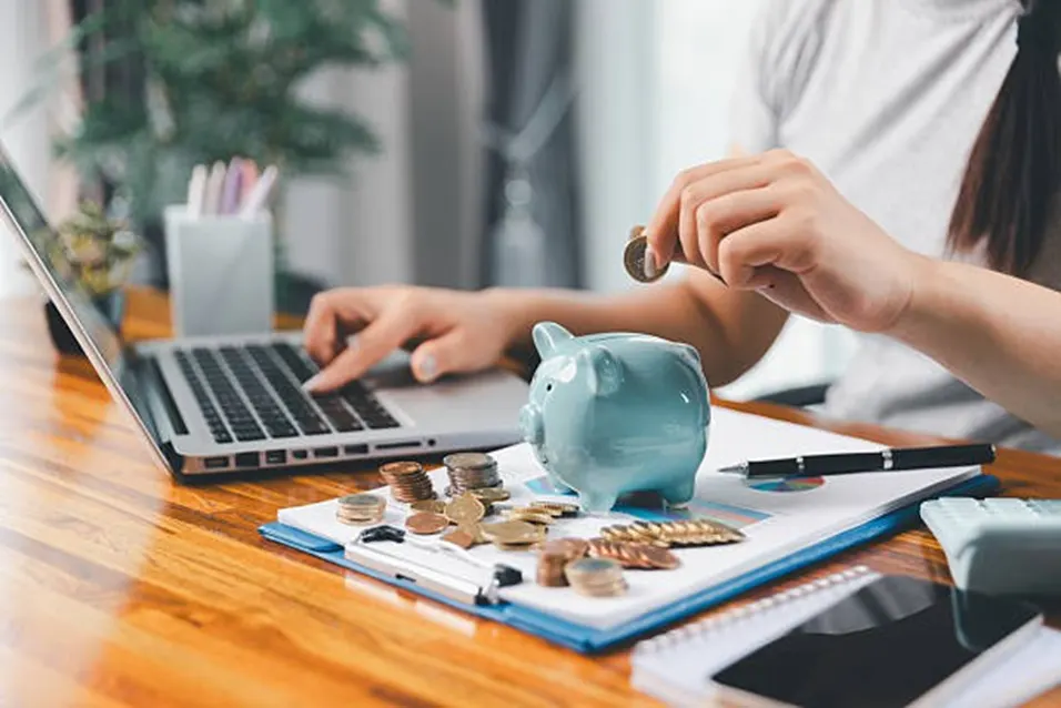 Woman inserting a coin into a piggy bank beside a laptop and financial charts