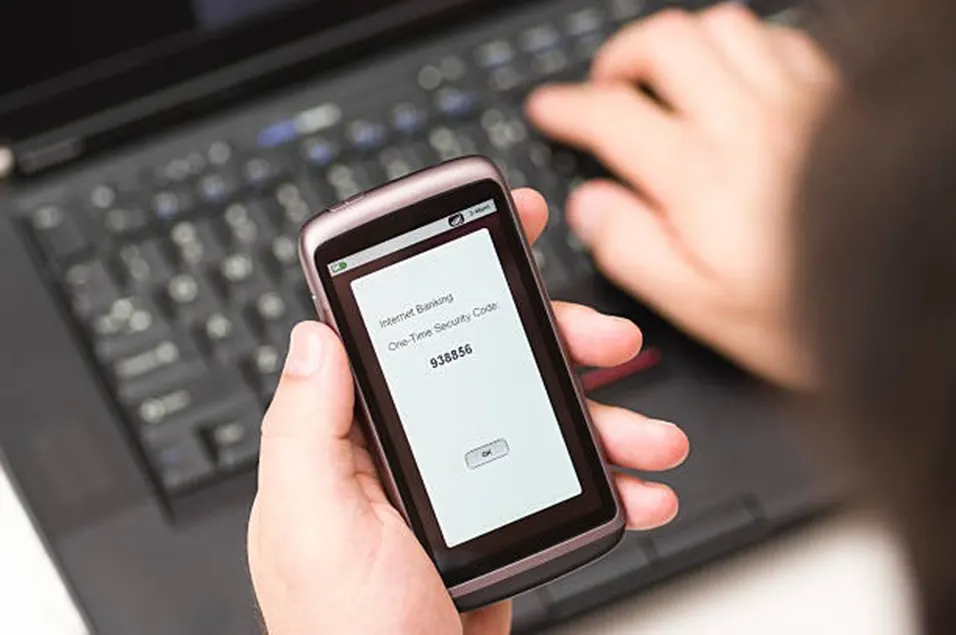 Woman holding a phone showing a one-time internet banking security code while typing on a laptop