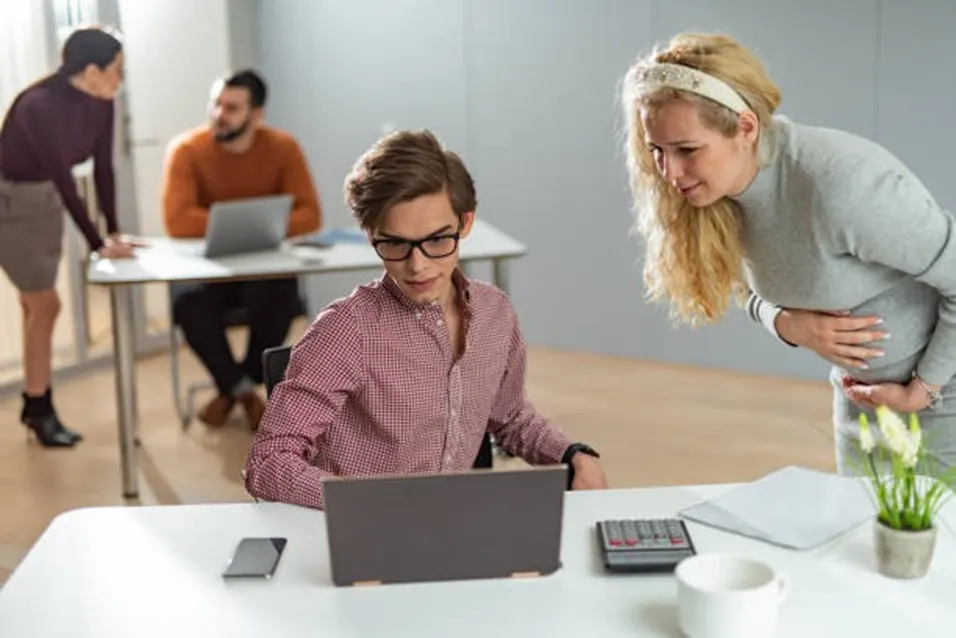 Pregnant woman in a grey jumper leaning over a colleague's laptop in a modern office with other colleagues in the background