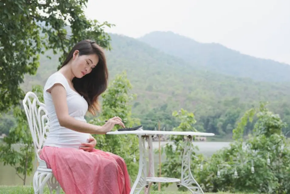 Pregnant woman in a white top and pink skirt sitting outdoors at a white garden table using a calculator with a green hillside behind her