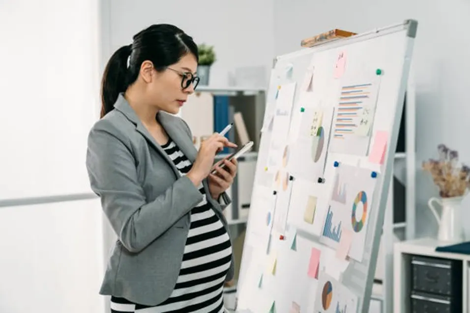 Pregnant businesswoman in a grey blazer and glasses reviewing a phone beside a whiteboard covered in charts and sticky notes