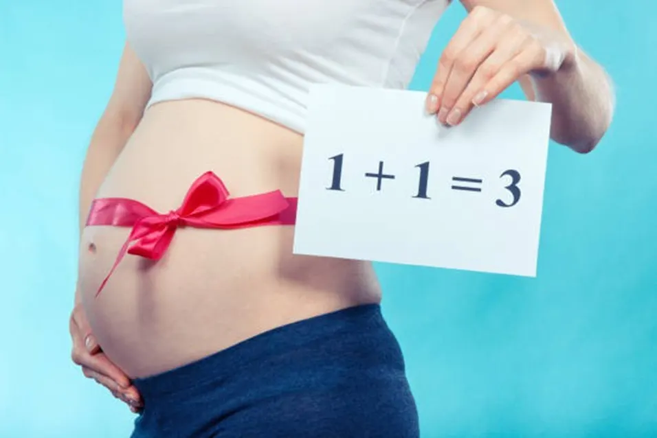 Pregnant woman holding a card showing 1+1=3 with a pink bow on her bump
