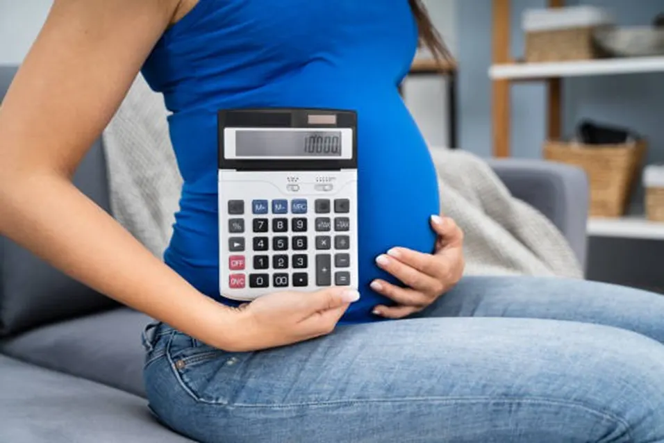 Pregnant woman in blue top holding a large calculator while seated on a sofa