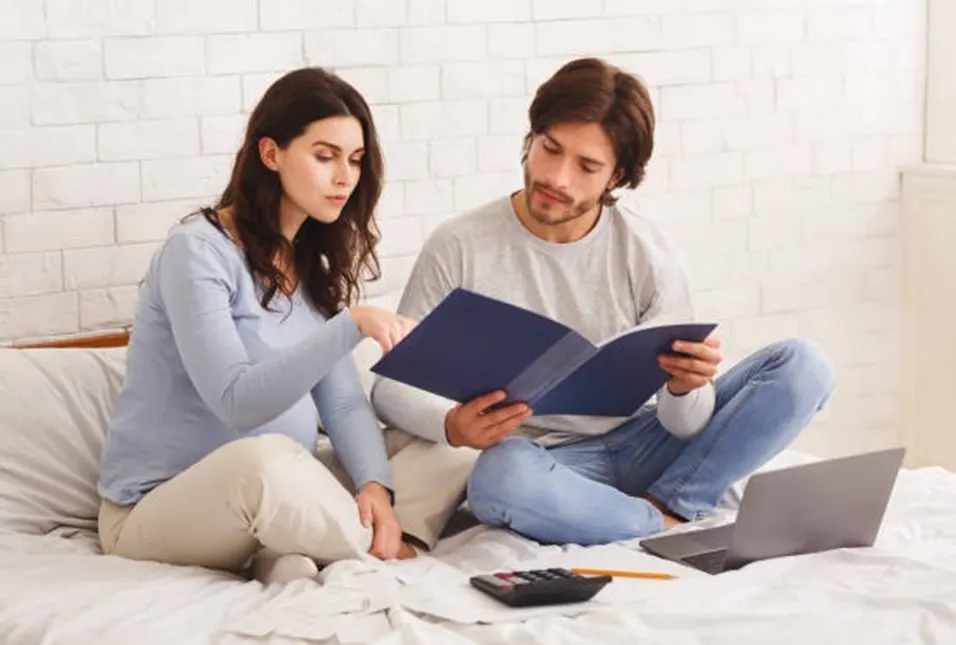 Pregnant woman and her partner sitting on a bed reviewing documents together with a calculator and laptop nearby