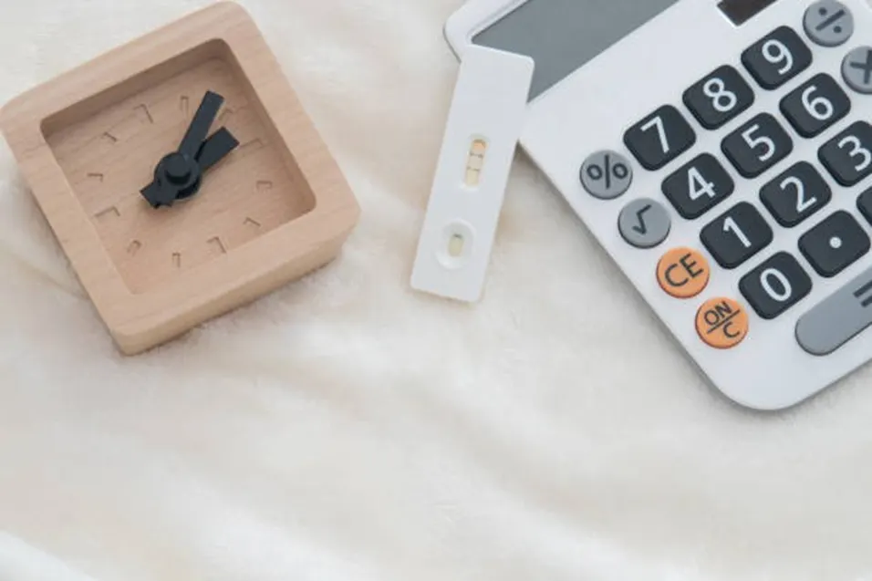 Wooden alarm clock, pregnancy test strip and white calculator laid out on a soft white surface