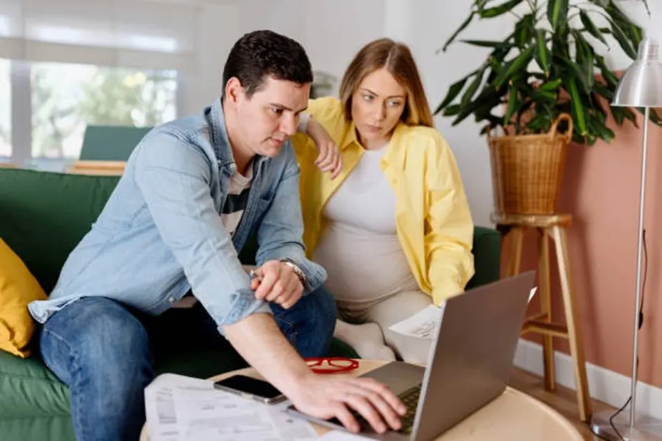 Man and pregnant woman sitting on a green sofa together looking at a laptop with documents and glasses on the table