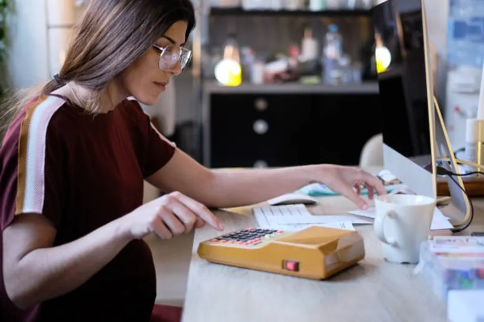 Young woman with glasses using a vintage yellow calculator at a desk with a computer monitor and mug nearby