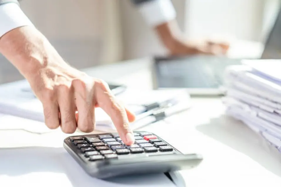 Person pressing calculator buttons at a bright office desk with documents and laptop in background