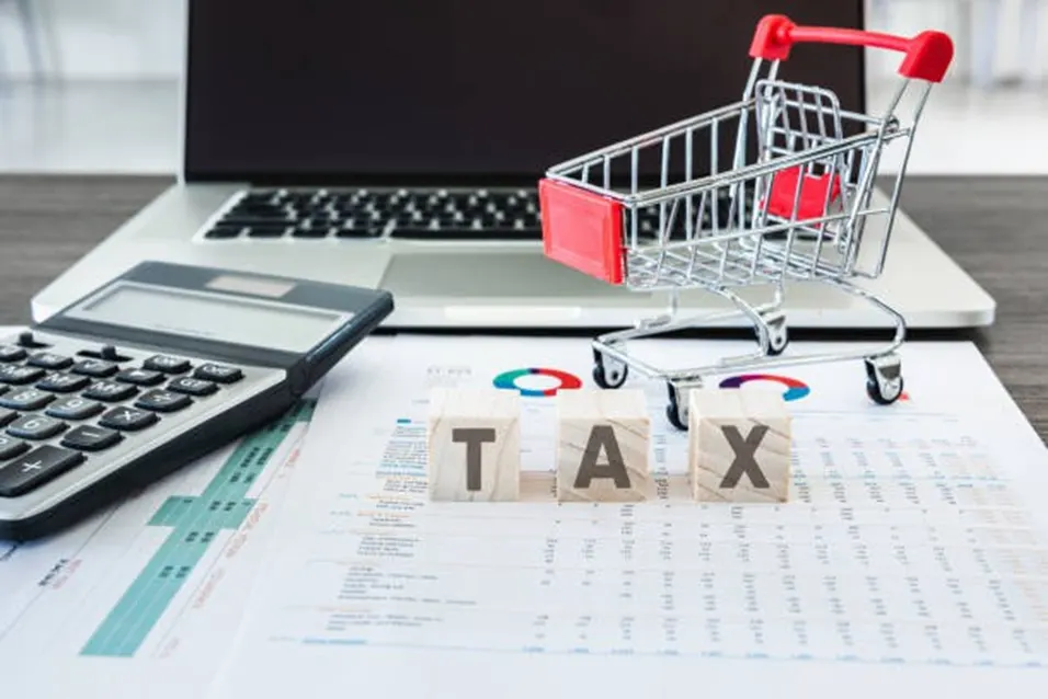 TAX wooden blocks beside a mini shopping cart and calculator on financial documents with a laptop