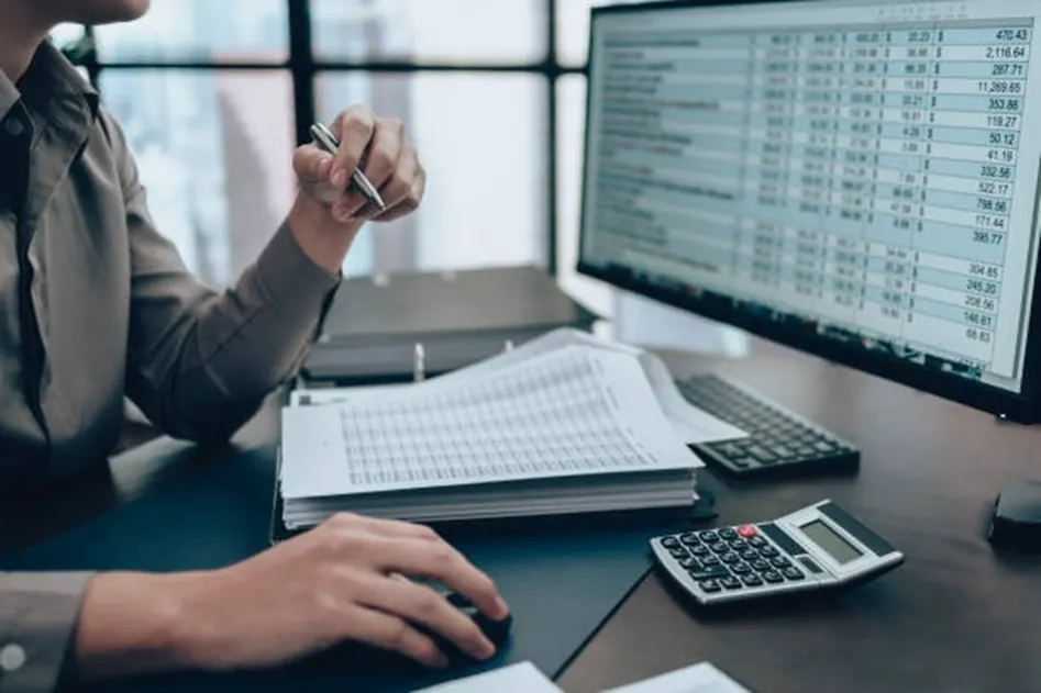 Accountant working at a desk with two calculators, financial reports, and a large monitor showing data