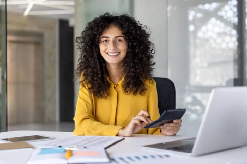 Smiling woman with curly hair in a yellow blouse holding a calculator at a bright office desk with a laptop