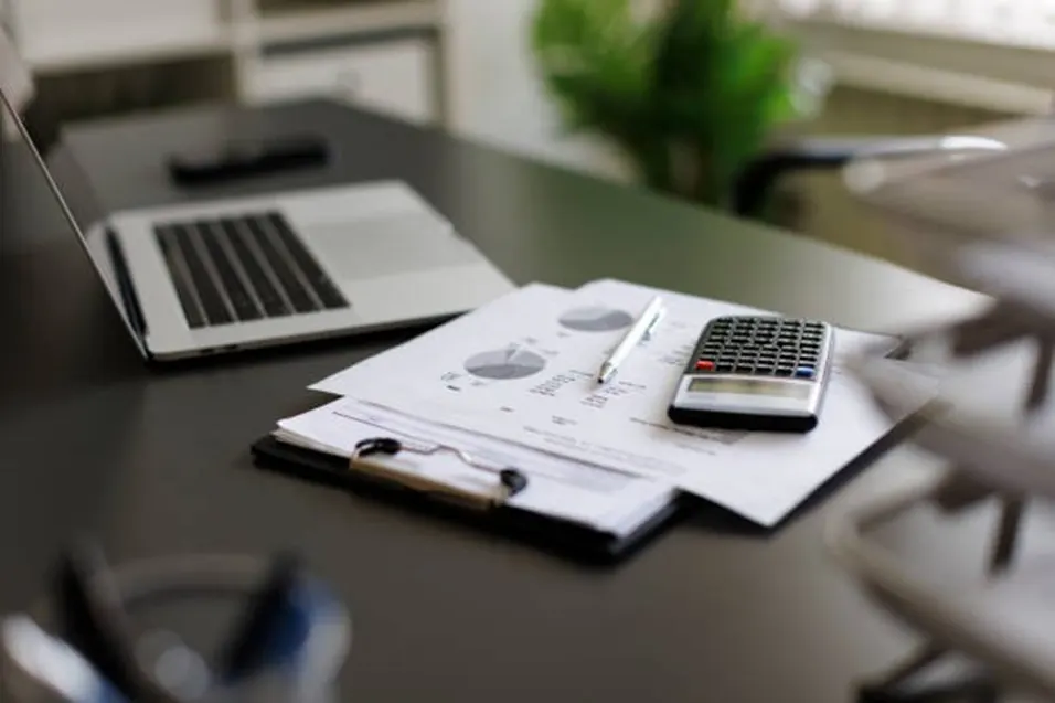 Calculator and pen resting on printed charts on a clipboard beside a laptop on a dark desk with a plant