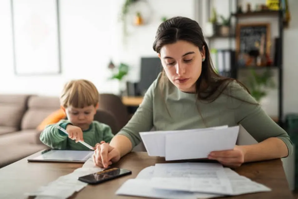 Mother reviewing documents and using a phone calculator at a table while her young child plays nearby