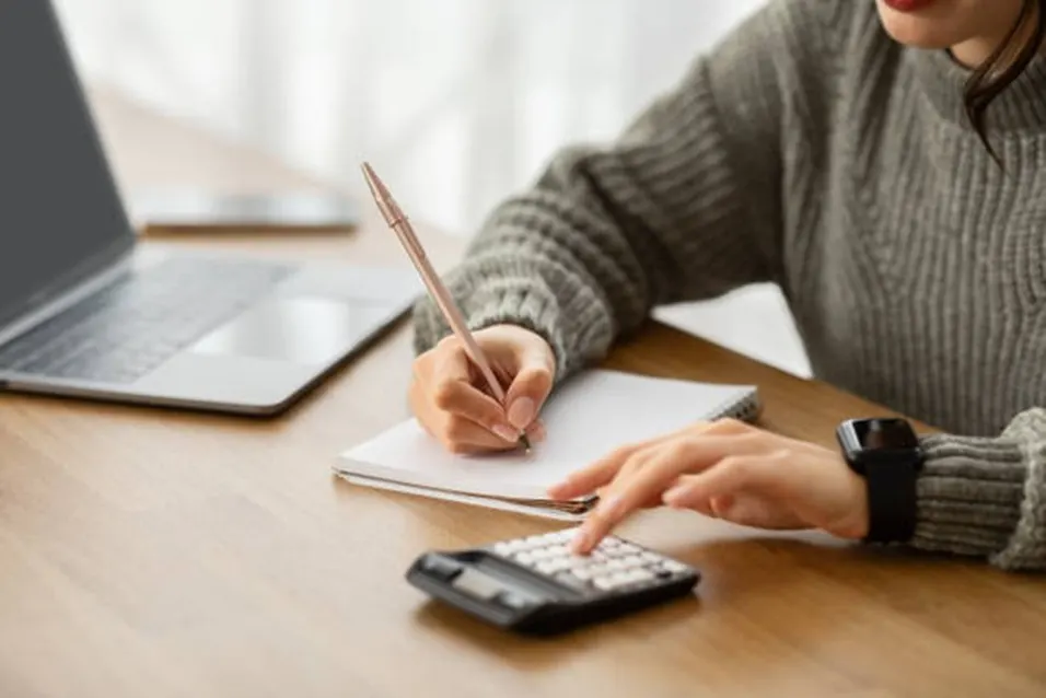 Woman in a grey knit sweater writing notes in a notebook while pressing a calculator beside a laptop