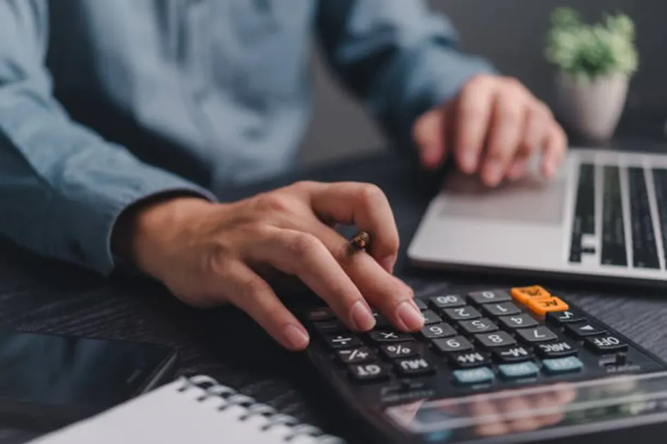 Person pressing buttons on a calculator with an orange key while using a laptop at a dark desk
