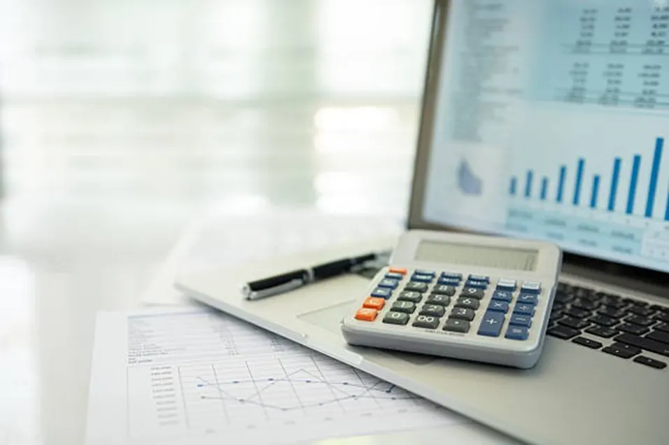 White and blue calculator resting on a laptop keyboard beside a pen and financial charts