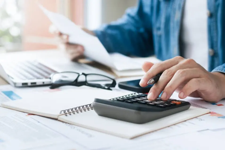 Person in denim shirt using a black calculator with a pen while reviewing documents and a laptop