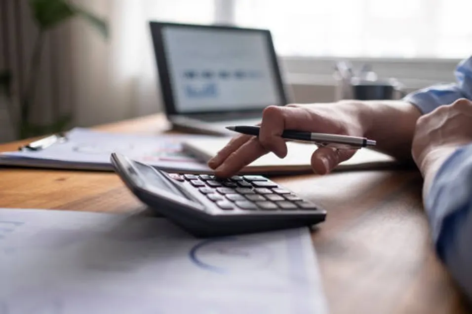Hand holding a pen while pressing a black calculator on a wooden desk with a laptop and charts