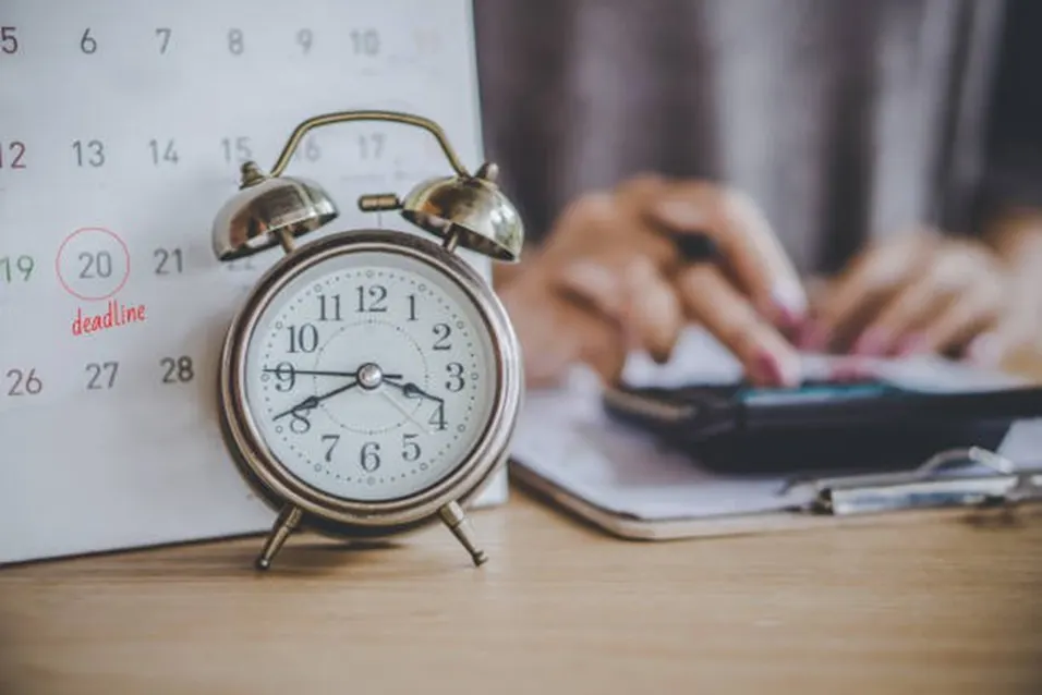 Vintage alarm clock in front of a calendar with a deadline circled, beside a person using a calculator on a wooden desk