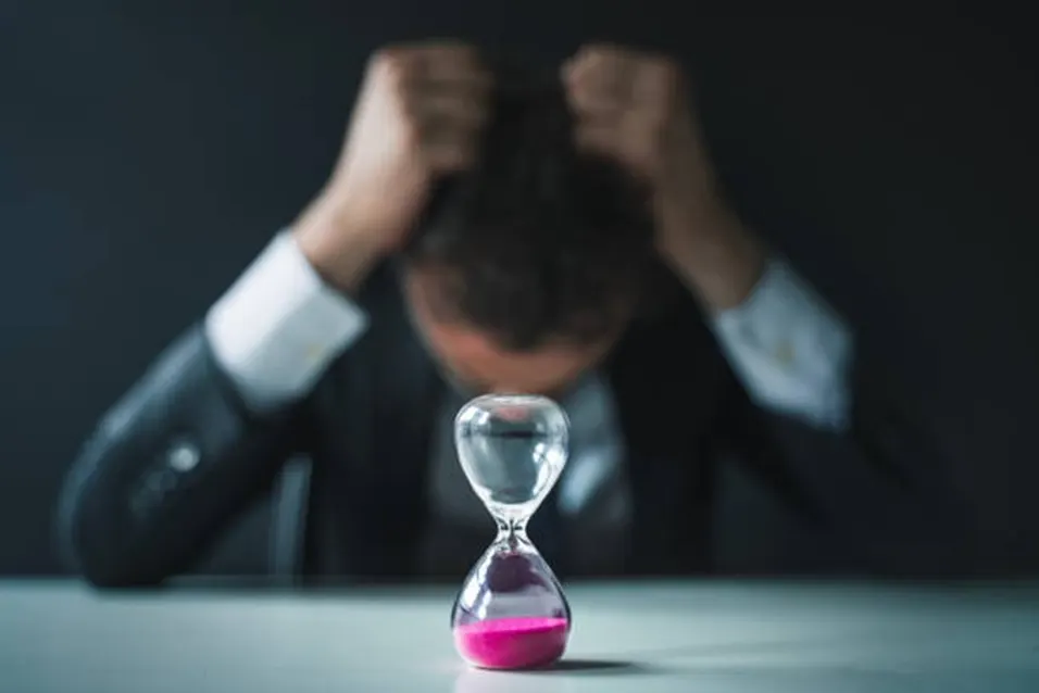 Man in a dark suit with head in hands, staring at an hourglass with pink sand running out on a dark background