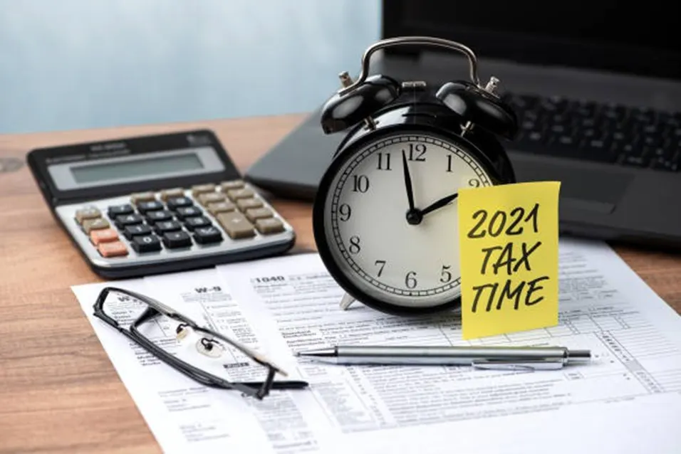 Black alarm clock beside a sticky note saying 2021 Tax Time, with a calculator, glasses and tax forms on a wooden desk